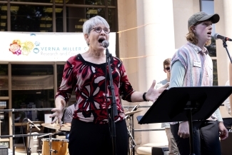 an older woman and a younger woman sing into microphones on an outdoor stage on a sunny day