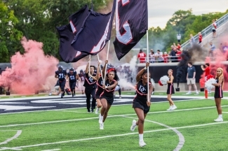 a group of young people in cheer uniforms run onto football field with flags and colorful smoke