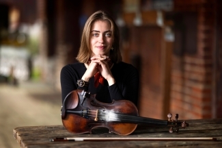 a young white woman with brown hair stands behind her fiddle in a rural setting