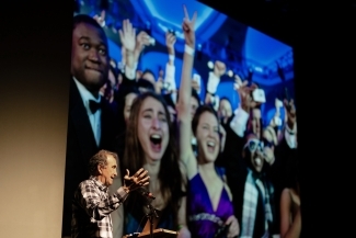 an older white man stands in front of a screen of photographs while speaking on stage