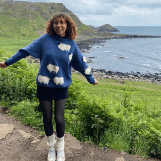 a young woman wearing a sheep sweater standing along a cliff-lined coastline
