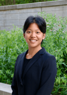 a young person with dark hair wearing a blazer in front of greenery