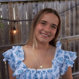 a young woman with blonde hair smiling in front of a wooden fence with string lights