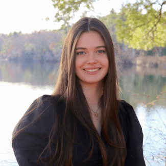 a young woman with long dark hair wearing a black top in front of a still lake on a sunny day
