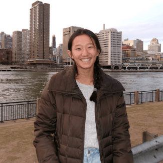 a young woman wearing a brown puffer jacket standing in front of a city skyline