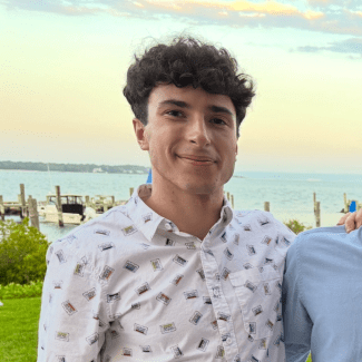 a young man with curly brown hair wearing a collared shirt in front of the ocean at sunset
