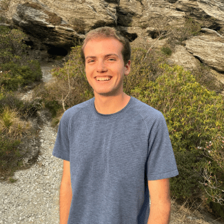 a young man with short blonde hair wearing a blue tshirt in front of a rocky landscape