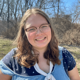 a young woman with curly brown hair wearing glasses outside on a sunny day
