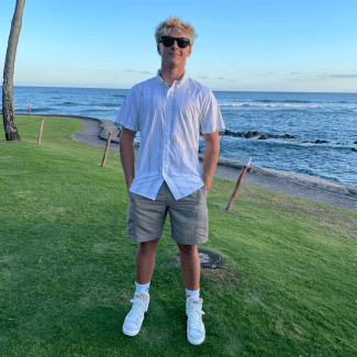 a young man with blonde hair wearing a white collared shirt and sunglasses near the ocean