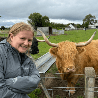 a young woman with blonde hair smiling next to a highland cow on a grey day