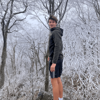 a young man with brown hair wearing a sweatshirt and shorts in front of a snowy setting