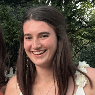 a young woman with dark hair wearing a white flowery top in front of a green landscape