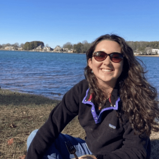 a young woman with long dark brown hair smiling with sunglasses in front of a river or lake