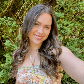 a young woman with curly brown hair smiling in front of a green bush