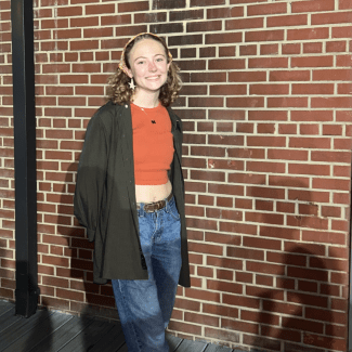 a young woman with curly blonde hari wearing a bandana and orange top in front of a brick wall