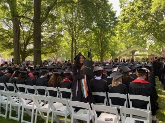 a young woman in a graduation cap and gown smiles and holds up a diploma on a sunny day outside