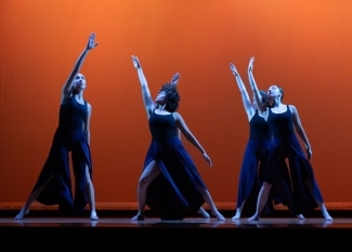 Four dancers in flowy costume are illuminated by blue stage lights against a contrasting orange background