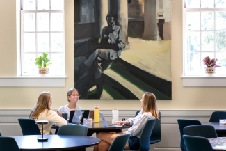 three young women smile while gathered around a table with laptops and waterbottles