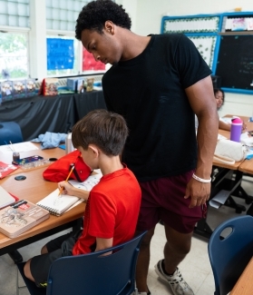 a young man assists a boy who is drawing at a desk