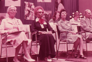 a Black woman sits in a row of desks alongside other white women and a man