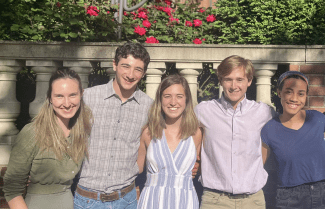 Students smiling, with arms around each other, in front of a rose bush at a Catholic church