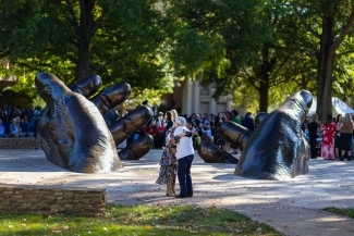 People hugging in front of a stately bronze sculpture of hands emerging from the earth