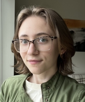 A college age person with shoulder-length light brown hair and glasses takes a selfie