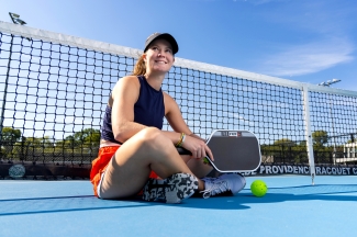a young woman sits on a pickleball court smiling and holding a paddle