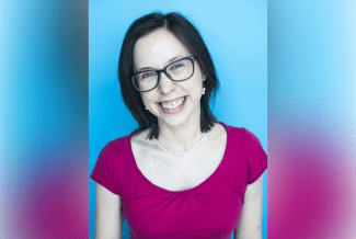 A smiling woman with shoulder-length brown hair and glasses, wearing a magenta shirt and a pearl necklace, against a solid bright blue background.