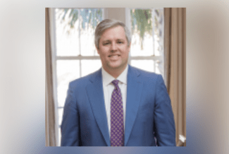 A professional headshot of a smiling man with short grey hair, wearing a blue suit, white shirt, and a patterned purple tie, positioned in front of a window with natural light.