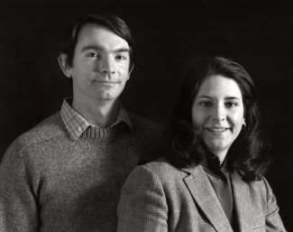 A black and white studio portrait of a man in a crewneck sweater and collared shirt standing next to a smiling woman in a patterned blazer.