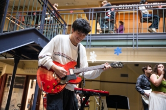 A smiling young man in a patterned sweater plays a red hollow-body electric guitar during a live performance in a multi-level indoor space.