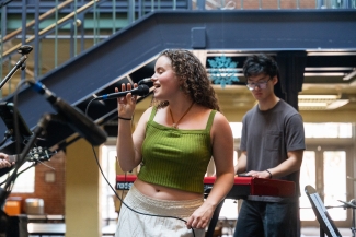 A woman with curly hair sings into a microphone on a stage while a man plays a keyboard in the background.