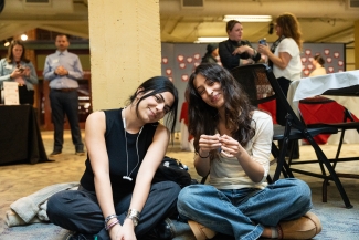 wo young women sit cross-legged on a carpeted floor, smiling toward the camera in a large event space.