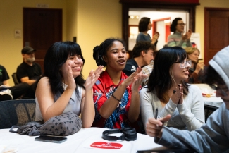 Three young women sit at a table with white linens, smiling and clapping their hands during an indoor event.