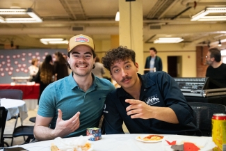 Two young men sit at a table together, one wearing a trucker hat and the other gesturing toward the camera during an event.