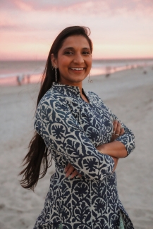 A smiling woman with long dark hair and crossed arms wearing a blue and white patterned tunic, standing on a sandy beach during a pink-hued sunset.