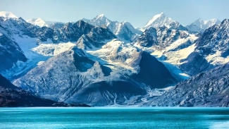 Snow-capped, rugged mountain peaks tower over a bright turquoise glacial bay under a clear blue sky.