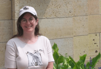 A smiling woman wearing a baseball cap and a shirt with a giraffe graphic stands in front of a stone wall and green plants.
