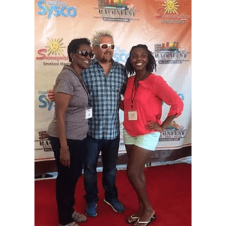 Guy Fieri stands on a red carpet between two women in front of a backdrop featuring Sysco and Baconfest logos.