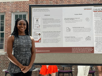A smiling young woman stands in front of a Davidson College research poster titled "2022-2023 FIRST Action Team Summary," which outlines initiatives for improving STEM culture and reframing neurodiversity in higher education.