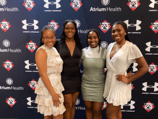 Four young women pose together and smile in front of a dark backdrop featuring repeated logos for Atrium Health, Under Armour, and a Davidson Wildcats mascot.