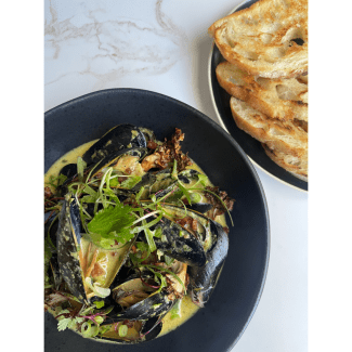 Steamed mussels in a creamy broth garnished with fresh herbs and microgreens, served with a side of toasted bread.