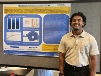 A smiling young man stands next to a research poster titled "Racism In COVID-19 and Biomedical Research," featuring data visualizations and sections on pulse oximetry and racial statistics in healthcare.