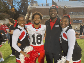 A Davidson College football player in a white and red jersey stands between two cheerleaders holding red and white pom-poms, alongside another man in a Davidson Football jacket.