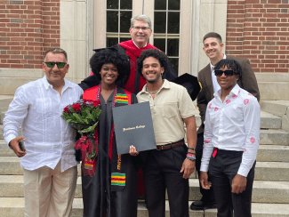 A group of six people, including a graduate in a black cap and gown holding a Davidson College diploma folder and flowers, pose together on brick stairs in front of a building.