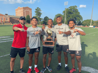 Five members of the Davidson Football team stand on an athletic field, proudly displaying championship rings and a trophy that reads "2020-21 CHAMPION."