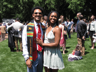 A smiling young couple poses on a sunny lawn during a graduation event; the man wears a graduation stole and tie, while the woman wears a light-colored summer dress.