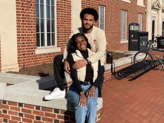 A smiling young man and woman pose together while sitting on a low brick wall in front of a brick campus building with arched windows.