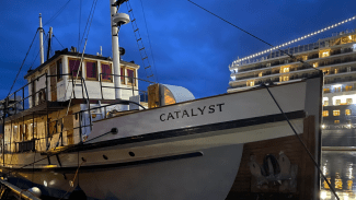 The wooden boat Catalyst is docked at night with glowing interior lights, while a large, multi-deck cruise ship illuminated with strings of lights sits in the background.